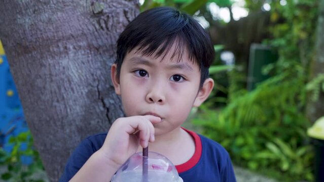 4K, Asian boy close up, sitting in park with playground, Sitting drinking water, Fatigued, drank pipo yoghurt smoothie deliciously, His hand grasped  water tube, looked friend playing in playground.
