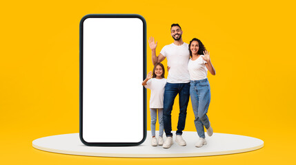Glad young middle-eastern family with small girl waving hands near huge phone with blank screen