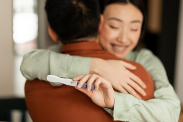 Obraz na płótnie Canvas Happy young asian woman holding positive pregnancy test and hugging husband, loving family embracing at home, closeup