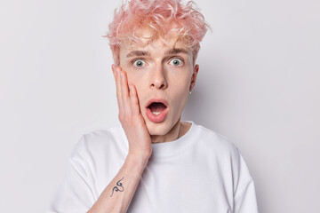 Shocked scared guy with pink hair looks with terror keeps hand on cheekk mouth widely opened reacts to scarying news has tattoo on arm dressed in t shirt isolated on white background. Human reactions