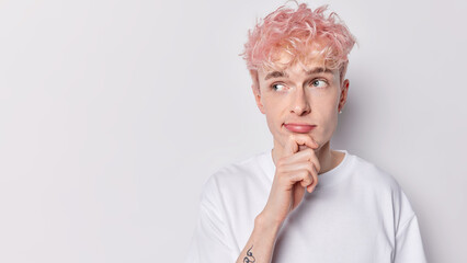 Horizontal shot of thoughtful pink haired man keeps hand on chin looks pensively aside has pensive expression dressed in casual t shirt isolated over white background blank space for your advert