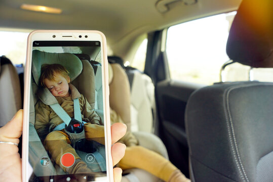 A woman takes a picture on her phone of her child, who fell asleep in the car in a child seat