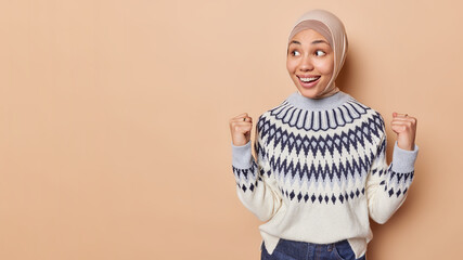 Joyful Arabic woman in headscarf and jumper clenches fists raises hands as gets great bonus smiles broadly focused aside isolated over brown background with copy space on left side for your advert