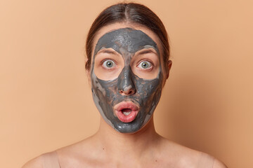 Headshot of surprised brunette young woman applies facial mud mask on face undergoes skin vitality procedures stares shocked at camera keeps mouth widely opened isolated over brown background