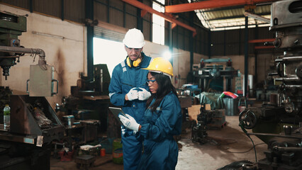 Two multiracial factory workers in safety wear are using a tablet to plan for working at a metalwork production factory. Male and female production engineers are examining the status of manufacturing.