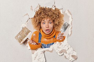 Scared curly female plasterer busy repairing wall in apartment holds brush and spatula being gypsum...