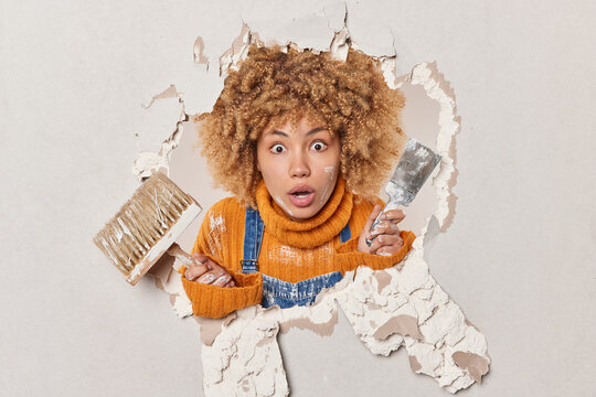 Scared curly female plasterer busy repairing wall in apartment holds brush and spatula being gypsum master holds breath from amazement poses in hole wears orange jumper. Home renovating concept