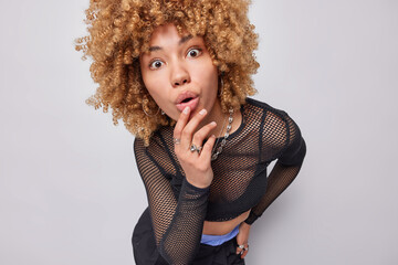 Photo of surprised curly haired woman keeps mouth widely opened stares with wonder at camera...