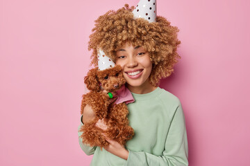 Portrait of cheerful curly haired young female model celebrates pets birthday wears party cone hat poses with small poodle puppy wears green pullover being on party isolated over pink background