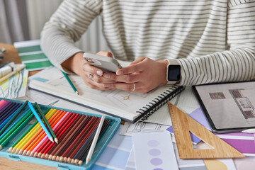 Cropped image of unrecognizable woman wears striped jumper and smartwatch holds smartphone sends messages or searches new ideas for interior design poses at table with crayons album with drawings