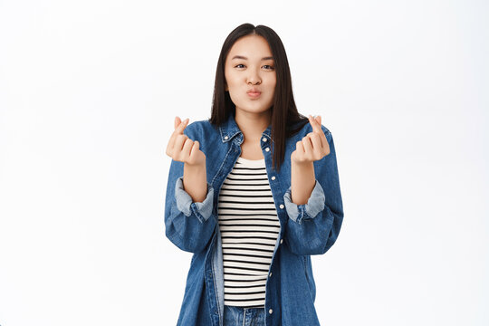 Cute young woman, 20 years old, asian girl shows heart finger sign, I love you gestures, stands over white background