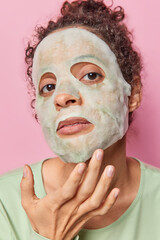 Fototapeta premium Headshot of serious woman applies moisturising sheet mask does skin care procedures touches face looks attentively at camera isolated over pink background background. Beauty procedures concept