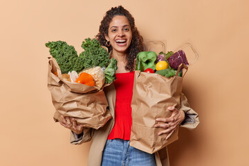 Horizontal shot of curly haired woman carries paper grocery bag full of vegetables returns from market smiles broadly has happy mood dressed in stylish clothes isolated over brown background