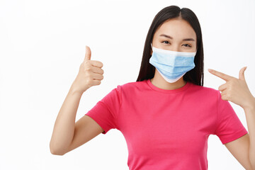 Asian smiling girl supports wearing medical face mask during covid-19, shows thumbs up, stands over white background