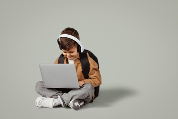 Busy cheerful caucasian 6 years old little boy in wireless headphones sits on floor, typing on laptop