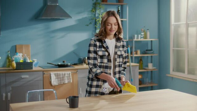 A Smiling Young Woman Housewife Holding a Spray Bottle and Cleaning a Table in the Kitchen with a Yellow Microfiber Cloth. Multi-functional Cleaning Tools.