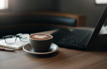 Close-up view, white cup of coffee with computer laptop, notebook, pen and eye glasses on wooden table in cafe, People resting after work in a calm cafe