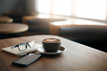 Fototapeta Close-up view, white cup of coffee with smartphone, notebook, pen and eye glasses on wooden table in cafe. Vintage light, blurred background obraz