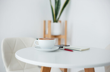 Minimal office workspace interior design. white coffee cup with mobile phone and note book on white table against white wall