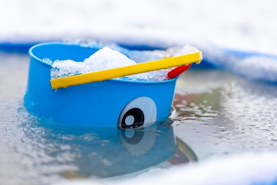 A close up portrait of a blue play bucket with a face painted on it and a yellow handle lying in a blue play shell pool which is frozen and surrounded by snow in the winter.