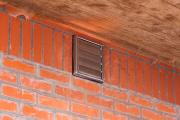 A portrait of a square brown design vent with closed slats placed in a red brick wall on a building for the air output of a cooker hood. all the fumes when cooking food are transferred outside.