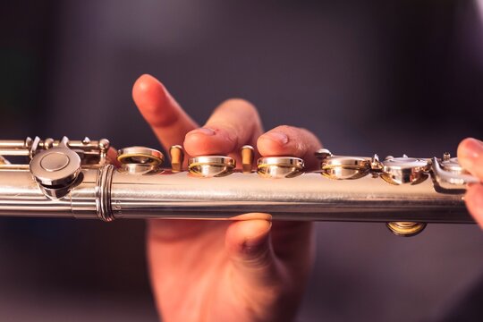 A portrait of the fingers of a hand of a flutist musician pressing down on the valves of a metal silver flute to play a note of a musical piece during a concert.