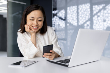 Fototapeta na wymiar Asian business woman smiling and working at the workplace inside the office, businesswoman reading news using smartphone, boss with laptop using phone and browsing internet pages online.