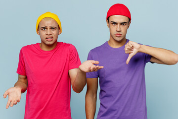 Young confused couple two friends men wear casual clothes together shrugging shoulders looking puzzled spread hands show thumb down dislike gesture isolated on pastel plain light blue cyan background.