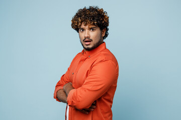 Side view young mad sad Indian man wearing orange red shirt white t-shirt hold hands crossed folded look camera isolated on plain pastel light blue cyan background studio portrait. Lifestyle concept.
