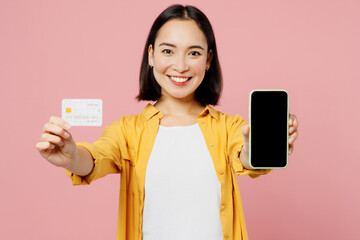 Naklejka premium Young woman of Asian ethnicity wear yellow shirt white t-shirt using blank screen mobile cell phone hold credit bank card shopping online order delivery booking tour isolated on plain pink background.