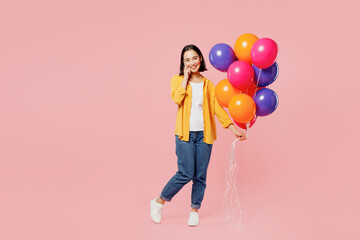 Young woman of Asian ethnicity wear yellow shirt white t-shirt hold face bunch of colorful air balloons look camera isolated on plain pastel light pink background studio portrait. Lifestyle concept.