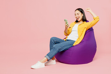 Full body young woman of Asian ethnicity wears yellow shirt white t-shirt sit in bag chair hold in...