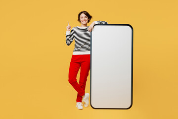 Full body young woman wear casual black and white shirt big huge blank screen mobile cell phone smartphone with workspace mockup area point index finger up isolated on plain yellow color background.