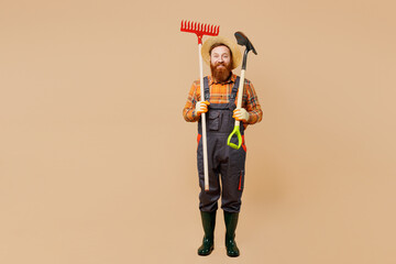 Full body fun young bearded man wear straw hat overalls work in garden hold in hand shovel rake...