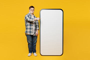 Full body young man with down syndrome wear glasses casual clothes point on big huge blank screen mobile cell phone isolated on pastel plain yellow color background. Genetic disease world day concept.