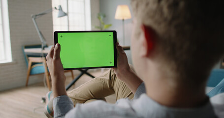 Over the Shoulder. Creative Young Caucasian guy Sitting at Sofa having an online video conference, using tablet with mock up green screen, doing distant learning during self-isolation