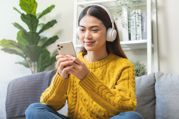 Relaxing asian young woman using smart phone listening to music, enjoy song or watching videos, podcast on mobile with white wireless headphones sitting on sofa, couch at home, chill out and leisure.