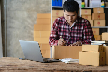 Asian Male SME Small Business Entrepreneur, freelance working from home using laptop for commercial auditing. online marketing Packaging Box SME Seller Concept Ecommerce Team online sales