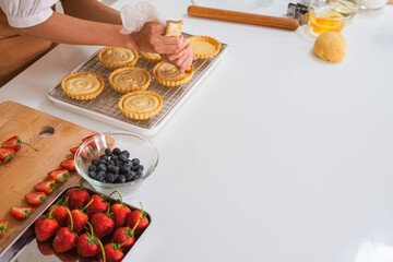 Young beautiful woman or housewife in apron standing at table with ingredients and baking tools in the kitchen at home making dessert by squeezing cream from pastry bag into freshly baked tarts.