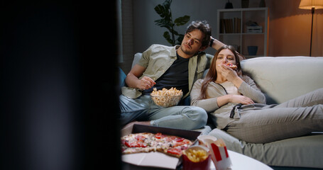 Caucasian extremely frustrated couple sitting in front of tv watching shows and eating pizza in...