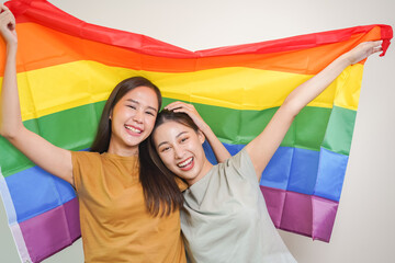 Happy lesbian, beautiful asian young two women, girl gay, couple love moment spending good time together, holding or waving lgbt rainbow, pride flag on isolated white wall background together at home.