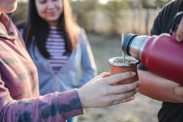  Close up of smiling friends drinking yerba mate using a thermos with hot water in the countryside...