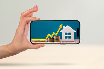 Fototapeta na wymiar Real estate market, graph, up arrow. House model and stack of coins. Hand with phone and copy space. On the screen of the phone the concept of cost, insurance, real estate mortgage.
