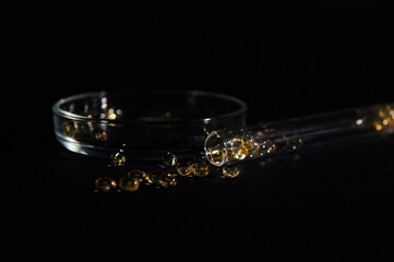Still life with soft translucent gelatin capsules of golden color with essential oils, omega-3 or fat-soluble vitamins scattered from a laboratory test tube, against a Petri dish on black background