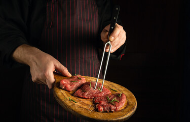 Chef holds a fork in his hand and a cutting board with beef steaks before barbecue. Dark space for restaurant recipe or hotel menu