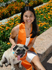 Young attractive Asian American college student, wearing gown and sash, celebrating her graduation from the university with here pug puppy dog