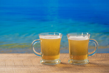 Two glasses of beer on a beach on wooden table with the sea water background on a sunny summer day, closeup