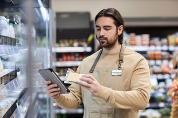 Waist up portrait of male worker in supermarket checking dairy product prices and expiry date