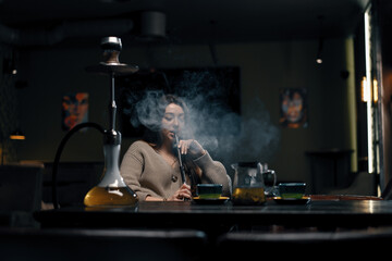 Beautiful young woman inhaling hookah girl smoking hookah sitting in lounge bar concept of...