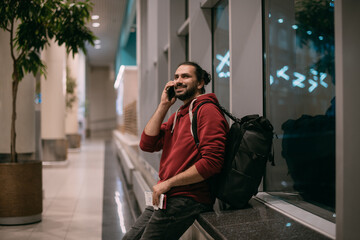 A  man is talking on the phone at the airport at night. A guy, a tourist with a  hand luggage, is waiting for departure at the gate
